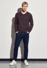 Mörklila V-ringad tröja, kombinerad med mörkblå jeans och vita sneakers. Modellen står med händerna i fickorna mot en enfärgad bakgrund.