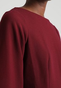 Viininpunainen, teksturoidusta kankaasta valmistettu paita, jossa on neliömäinen pääntie ja leveät, pitkät hihat. Näkyvät saumakohdat ja sileä, jäsennelty ulkonäkö.