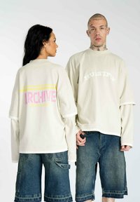 Cremfarbene, oversized T-Shirts mit kontrastierenden Grafiken: "ARCHIVE" in Pink und Gelb, "MULTIPLY" in Weiß; kombiniert mit Jeansshorts.