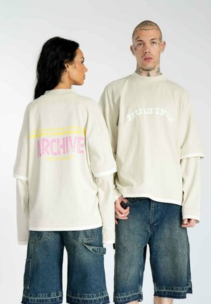 T-shirts oversize crème avec des graphiques contrastés : "ARCHIVE" en rose et jaune, "MULTIPLY" en blanc ; associés à un short en denim.