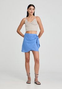 Top corto in maglia beige con spalline regolabili e gonna avvolgente blu legata di lato. La modella indossa anche sandali neri con cinturini.