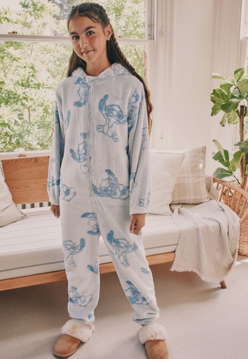 Fille portant une grenouillère à capuche bleu clair avec un motif de personnage de dessin animé et des chaussons duveteux, debout dans une pièce ensoleillée avec un banc en bois et des plantes.