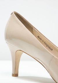 Chaussure à talon haut en cuir verni beige avec un bout pointu et un talon aiguille en bois fin. Étiquette indiquant "Fabriqué au Bangladesh."