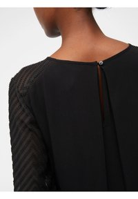 Vestido negro con mangas transparentes y estampadas que cuenta con un cierre de botón en la parte posterior y un sutil detalle de abertura, resaltando una textura suave.