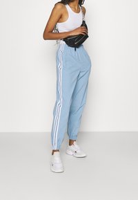 Kvinna som bär ljusblå träningsbyxor med vita ränder, vit linne, vita sneakers och håller en svart midjerväska över bröstet.