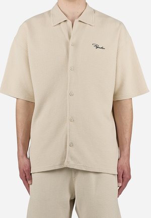 Chemise beige à manches courtes avec texture gaufrée, col et petit logo "Pegador" sur la poitrine gauche, portée avec un pantalon assorti.