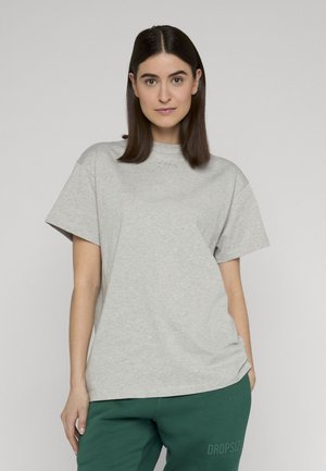 Žena s rovnými tmavými vlasy, oblečená v šedém oversized tričku a tmavě zelených kalhotách, stojící před jednobarevným pozadím.