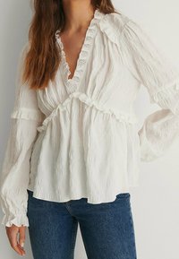 Blusa bianca a maniche lunghe con colletto e polsini arricciati, caratterizzata da un tessuto testurizzato e un design con vita arricciata. Abbinata a jeans blu.