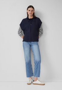 Marinefarbener Pullover mit Kordelzug am Kragen, der über einem gestreiften Langarmshirt getragen wird, kombiniert mit hellblauen Jeans und weißen Sneakers.