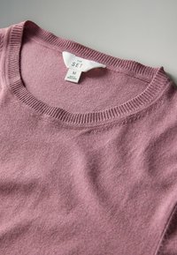 Maglione a maglia di un delicato viola chiaro con collo a costine e un'etichetta bianca con la scritta "THE SET M Made in Bangladesh" su una superficie grigia.