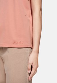 Blush pink T-shirt made of soft fabric, featuring subtle embellishments, side slits, and a logo stitched at the hem for detail.