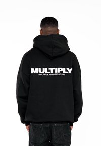 Sweat à capuche noir avec un grand texte blanc « MULTIPLY » au dos, doté d'une capuche à cordon de serrage et d'une coupe ample. Porté avec un jean foncé.