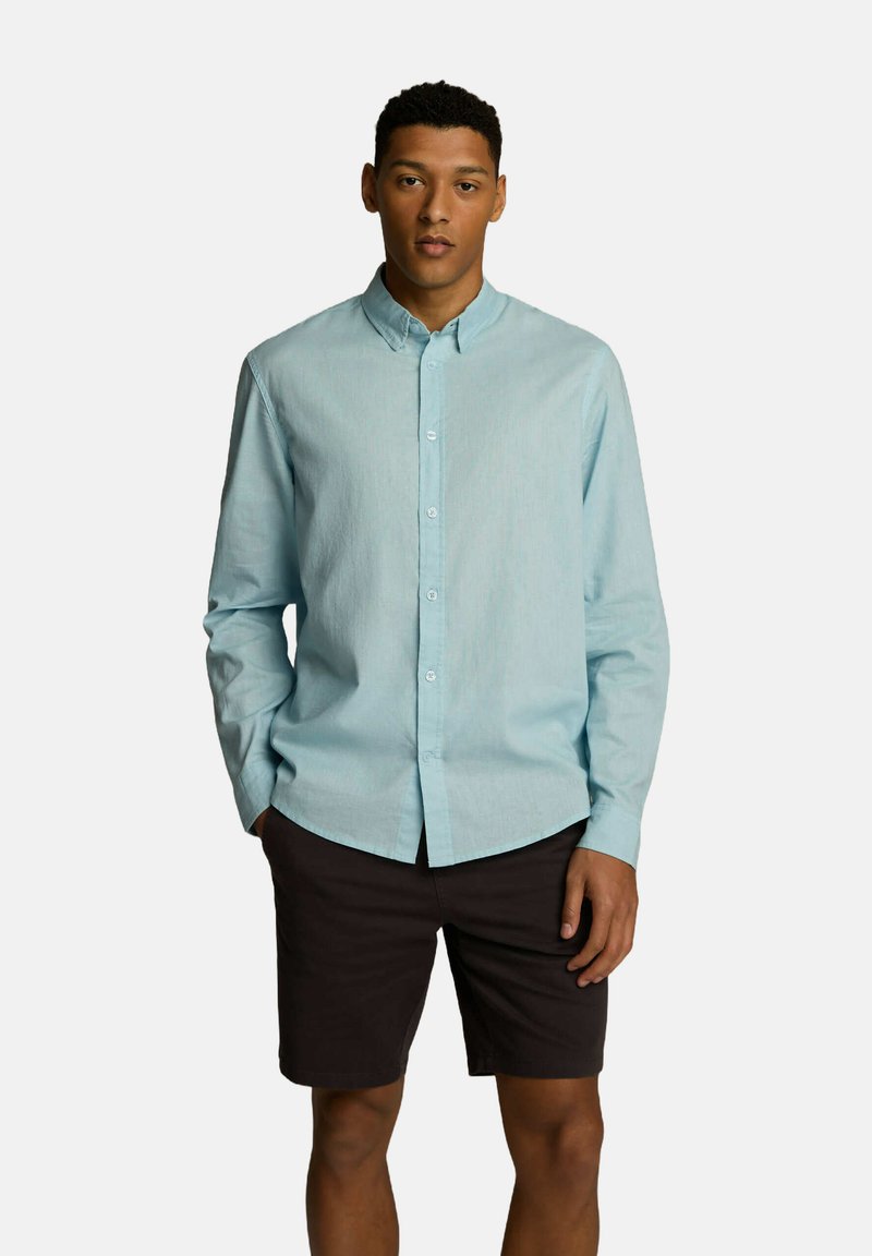 Jasnoniebieska koszula z długim rękawem wykonana z fakturowanej tkaniny, z kołnierzykiem i prostym dołem, zestawiona z ciemnymi szortami.