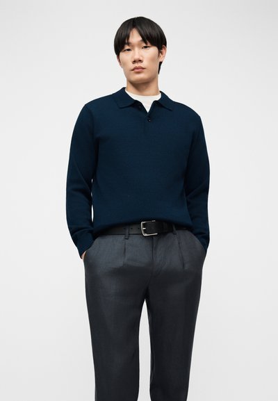 Homme portant un polo bleu marine à manches longues, un pantalon plissé gris avec ceinture noire, debout les mains dans les poches contre un fond uni.