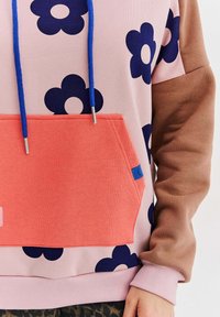 Mikina s kapucňou, ktorá má ružovú látku s tmavomodrými kvetinovými vzormi, hnedé rukávy a oranžovú prednú kombinovanú vrecko s modrými detailmi.