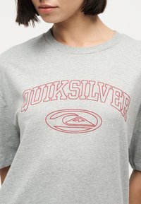 Γυναίκα που φοράει ανοιχτό γκρι μπλουζάκι με κόκκινο κείμενο "QUIKSILVER" και ένα στυλιζαρισμένο λογότυπο κύματος και βουνού στο στήθος.