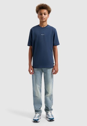 Tienerjongen staand, gekleed in een marineblauw T-shirt, lichtblauwe jeans en witte sneakers met blauwe accenten, tegen een effen grijze achtergrond.