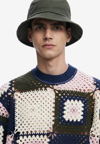 Häkelpullover mit mehrfarbigen Quadratmustern in Creme, Pink, Marineblau und Grün. Gerippter marineblauer Kragen. Olivgrüner Bucket Hat auf dem Modell.