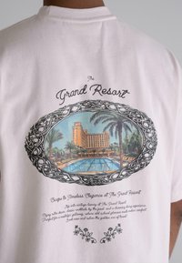 Light pink cotton t-shirt featuring an illustration of a resort with palm trees, a pool, and ornate decorative border on the back.