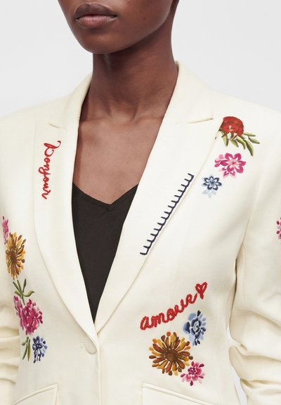 Femme portant un blazer crème avec une broderie florale colorée et les mots « bonjour » et « amour » en rouge sur les revers, sur un haut noir.