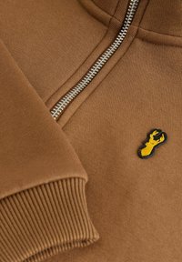 Hoodie zippé marron en tissu doux ; présente un poignet côtelé et un logo de homard jaune brodé sur fond noir. Détail de zip argenté.