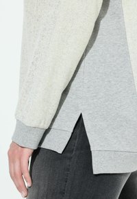 Primo piano di una persona che indossa una giacca beige con texture sopra una felpa grigia con spacchi laterali e pantaloni scuri, mostrando la mano sinistra rilassata.