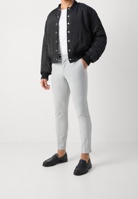 Person som bär en svart bomberjacka, vit skjorta, ljusgrå slim-fit byxor och svarta lädersandaler som står på en vit golv.