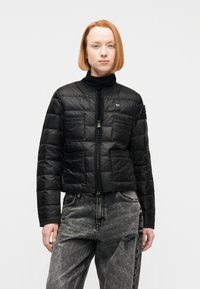 Γυναίκα με κοντά κόκκινα μαλλιά, φοράει μαύρο καπιτονέ μπουφάν και μαύρο τζιν ψηλής μέσης με φθαρμένο αποτέλεσμα και σκίσιμο στο αριστερό μηρό.