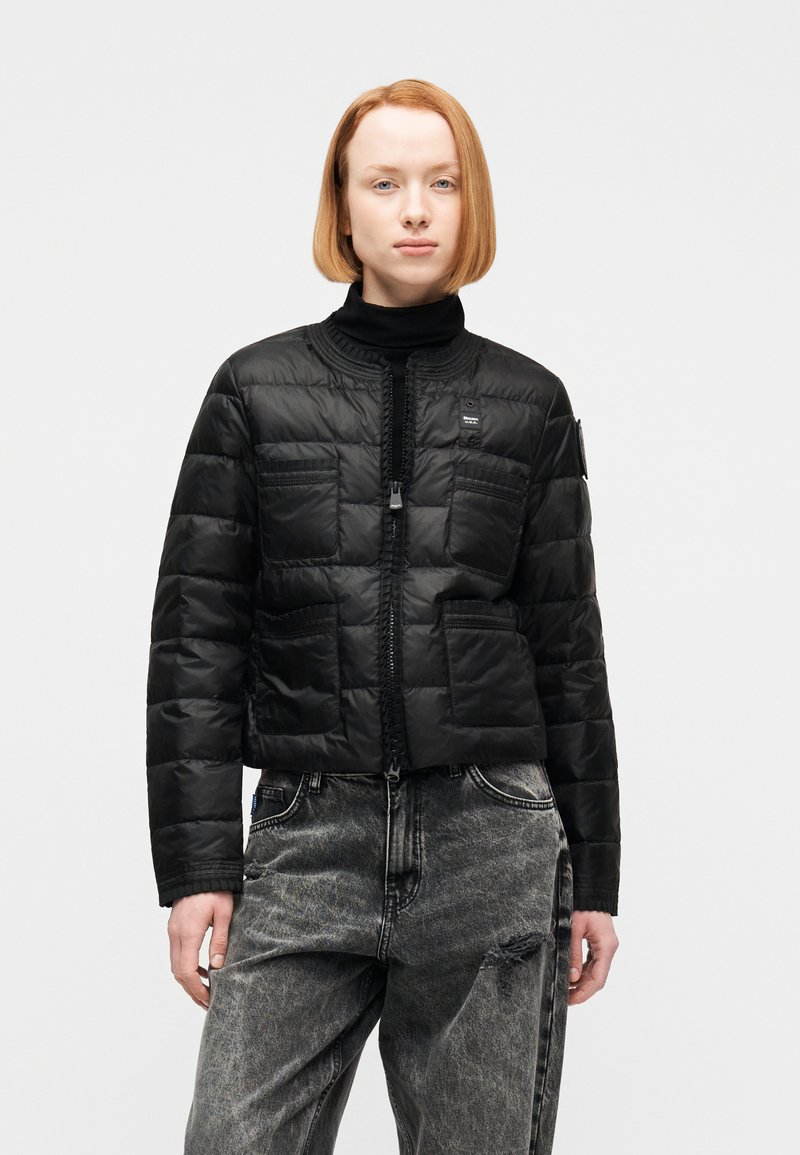 Γυναίκα με κοντά κόκκινα μαλλιά, φοράει μαύρο καπιτονέ μπουφάν και μαύρο τζιν ψηλής μέσης με φθαρμένο αποτέλεσμα και σκίσιμο στο αριστερό μηρό.