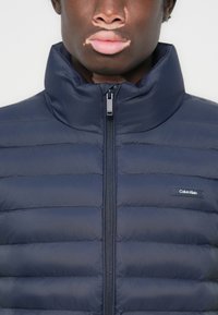 Navy padded jacket with high collar, full-length zipper, and horizontal quilted design. Features a small logo patch on the left side.