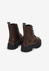 Botas de cuero marrón oscuro con cordones, suelas gruesas negras y cremalleras laterales, mostradas desde un ángulo trasero sobre un fondo blanco.