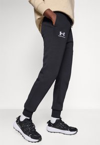 Čierne teplákové nohavice so bočnými vreckami a elastickými manžetami, s bielym logom Under Armour na stehne. Hladká textúra, pohodlný strih.