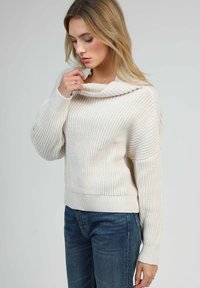 Pull en maille de couleur crème avec un col en cowl, texture côtelée et manches longues ; associé à un jean en denim bleu foncé.