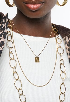 Gouden sieraden bevatten gelaagde kettingen, waaronder een met een rechthoekig label en een getextureerd ovaal-linkontwerp, gedragen over een witte geribbelde top.