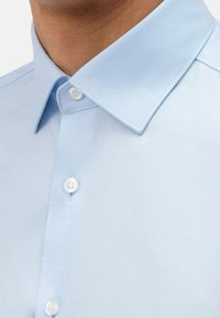 Chemise bleu clair boutonnée avec col structuré, texture lisse et lignes diagonales discrètes ; boutons blancs et arrondis.