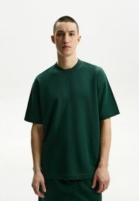 Sötétzöld, túlságosan nagy póló rövid ujjakkal és kerek nyakkivágással, sima textúrájú anyagból készült. Egyszerű dizájn, minta vagy kiemelkedő részletek nélkül.