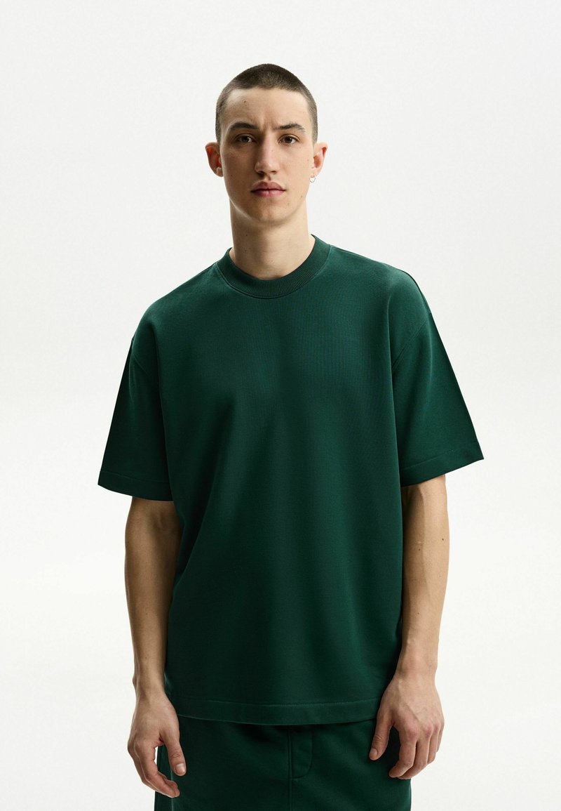 Sötétzöld, túlságosan nagy póló rövid ujjakkal és kerek nyakkivágással, sima textúrájú anyagból készült. Egyszerű dizájn, minta vagy kiemelkedő részletek nélkül.