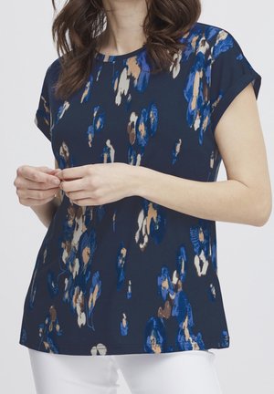 Donna che indossa una camicetta blu scuro a maniche corte con fantasia floreale astratta in blu, marrone e beige e pantaloni bianchi.