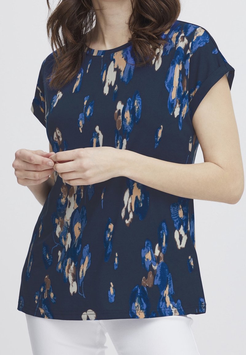Femme portant un chemisier bleu marine à manches courtes avec un motif floral abstrait bleu, marron et beige et un pantalon blanc.