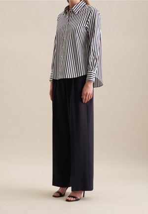 Kvinde iført sorte bukser med vide ben, sorte højhælede sandaler og en langærmet sort og hvid skjorte med lodrette striber.