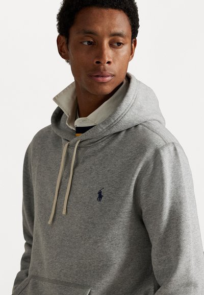 Giovane uomo con capelli ricci, che indossa un hoodie grigio sopra una camicia con colletto, mentre guarda di lato su uno sfondo bianco semplice.