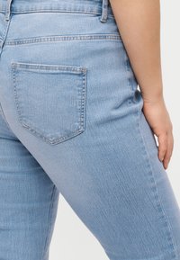 Κοντινό ενός ατόμου που φοράει ανοιχτό μπλε τζιν, εμφανίζοντας την πίσω τσέπη και μέρος του δεξιού βραχίονα που ακουμπάει δίπλα στο σώμα.