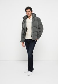 Chaqueta de plumas gris con costuras horizontales, capucha fija, cierre frontal y bolsillos laterales, usada sobre una camisa ligera y vaqueros.