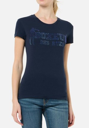 Donna che indossa una t-shirt aderente blu navy con testo scintillante "BOXEUR DES RUES" e sagoma di un pugile, abbinata a jeans blu.