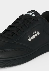 Μαύρο αθλητικό παπούτσι Diadora με μαύρα κορδόνια και λευκό λογότυπο της εταιρείας στο πλάι και στη γλώσσα, εμφανιζόμενο σε κοντινή λήψη με απλό φόντο.