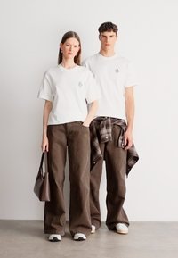 Fehér pamut pólók laza barna nadrággal párosítva. Az egyik modellnek kockás ing van derékban megkötve; mindketten sötét színű táskát visznek.