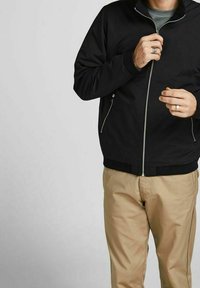 Veste noire à fermeture éclair avec un col ajusté, deux poches latérales et des poignets côtelés, portée sur un haut gris et associée à un pantalon beige.