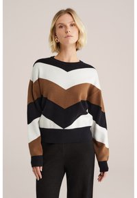 Blonda modele valkā garām piedurknēm, zigzaga raksta džemperi brūnā, baltā un melnā krāsā, saskaņojot ar tumšām biksēm, fona ir neitrāls.