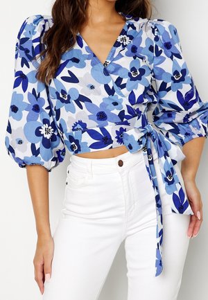 Kvinde iført en blå blomstret slå-om-bluse med puffede ærmer og hvide bukser med høj talje, hånd hvilende ved siden af sig.