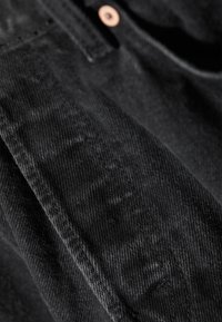 Gros plan sur un jean en denim noir montrant la texture du tissu, les coutures et un seul bouton en rivet de cuivre près de la ceinture.